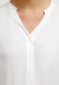Weißes, leichtes, knöpfbares Hemd mit V-Ausschnitt und weichem Textil, mit dezentem Glanz und klarem Design. Die Knöpfe sind rund und gleichmäßig angeordnet.