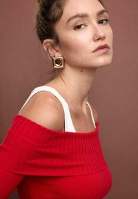 Roter schulterfreier, gerippter Pullover mit einem weißen Träger darunter. Das Modell trägt einen goldenen quadratischen Ohrring. Der Hintergrund ist in einem gedämpften Braunton gehalten.