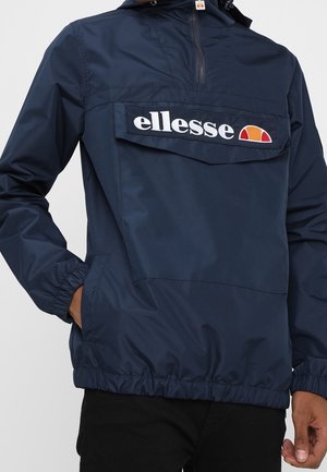 Navyblauer ellesse-Windbreaker mit Fronttaschenklappe und Logo, getragen von einer Person mit teilweise in den Taschen steckenden Händen.