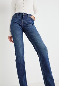 Modelis vilki aukšto liemens, tamsiai mėlynos tiesios kojos džinsus, viena ranka kišenėje, ir baltą ilgomis rankovėmis, suknelių marškinėlį su mygtukais.