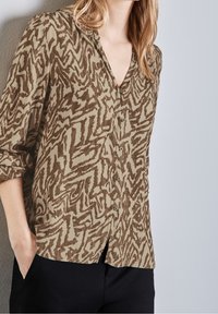 Femme portant une chemise boutonnée à motifs abstraits marron et beige, manches retroussées, associée à un pantalon noir, debout contre un mur clair.