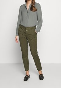 Blouse grise à manches longues avec fermeture à boutons, associée à un pantalon slim vert olive et des mocassins noirs ornés d'accents métalliques dorés.