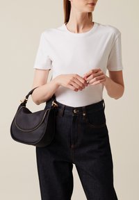 Borsa a tracolla in pelle nera con forma curva, superficie testurizzata, cerniera dorata e manico intrecciato corto, abbinata a una maglietta bianca semplice e jeans scuri.