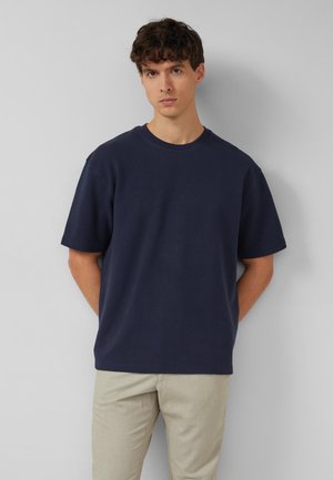 Jeune homme aux cheveux bouclés foncés, portant une chemise bleu marine à manches courtes et un pantalon beige clair, debout contre un mur clair uni.