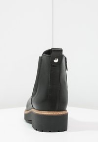 Stivaletto in pelle nera con suola spessa, con linguetta sul retro con bottone in metallo e zip laterale, posizionato su una superficie bianca contro un muro semplice.