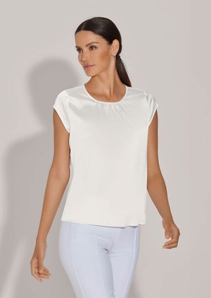 Woman with dark hair in a ponytail wearing a white sleeveless blouse and fitted white pants, looking to the side against a plain background.