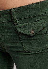 Pantalons en mélange de coton vert présentant une surface texturée, une petite poche à rabat avec un bouton en métal et des détails de couture précis.