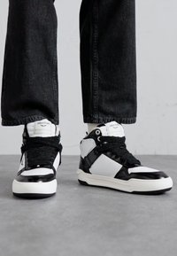 High-top sneakers in black and white, featuring a textured black strap, shiny black accents, and subtle logo on the tongue.