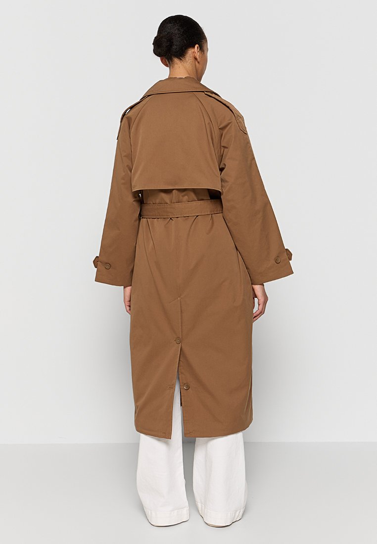 Manteau trench brun avec une taille cintrée et un col haut, présentant un design ceinturé, des accents de boutons et une fente arrière pour plus de mobilité.