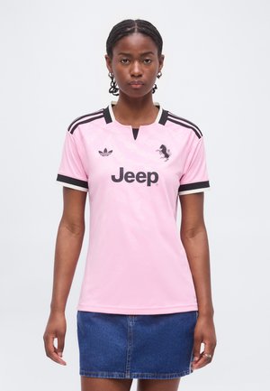 Joven mujer con cabello trenzado que lleva una camiseta deportiva rosa claro con rayas negras de Adidas y el logo de Jeep, combinada con una falda azul de mezclilla.