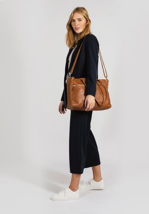 Brown leather shoulder bag with a structured design, adjustable strap, and two front zippered pockets. Model wears navy outfit and white sneakers.