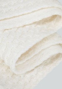 Coperta cremosa, soffice e strutturata con un motivo a maglia, caratterizzata da molteplici pieghe, realizzata in un tessuto accogliente ideale per calore e comfort.