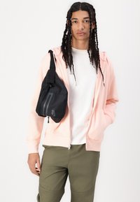 Mala de ombro preta com detalhes cinzentos, usada sobre um hoodie com fecho éclair rosa claro e uma t-shirt branca, combinada com calças verde-oliva.
