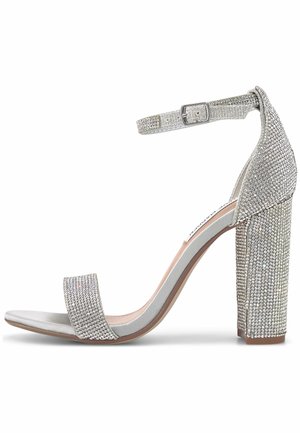 Silver high-heeled sandal featuring a textured strap and buckle. The block heel is adorned with rhinestones, creating a sparkling effect.