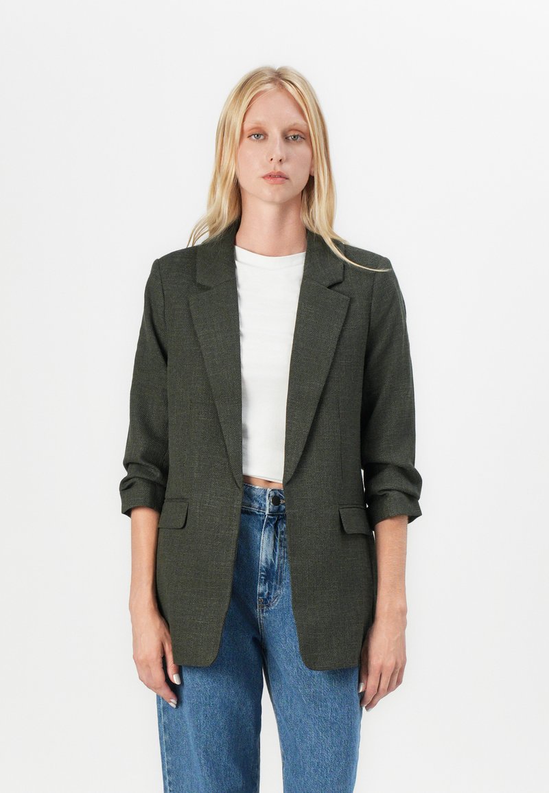 Tmavě zelený blazer s texturovanou tkaninou, jednořadým designem, klopovými límci a ohrnutými rukávy. Spárováno s džíny s vysokým pasem.