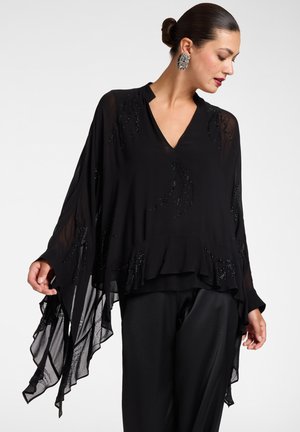 Donna che indossa una camicetta nera trasparente e ampia con dettagli di perline e maniche larghe e fluide abbinata a pantaloni neri, capelli raccolti in uno chignon e orecchini vistosi.