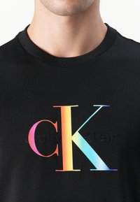 Juodas medvilnės marškinėliai su dideliu daugiaspalviu "CK" logotipu ir iškilniais "Calvin Klein" tekstu po juo. Paprasto apvalaus kaklo dizainas.