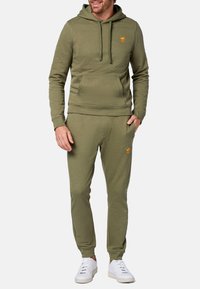 Set di felpa e pantaloni jogger verde oliva, con tasca a marsupio, polsini a coste e un logo arancione sul petto e sull'anca sinistra. Sneakers bianche.