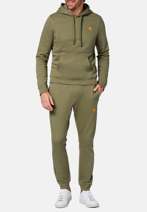 Set di felpa e pantaloni jogger verde oliva, con tasca a marsupio, polsini a coste e un logo arancione sul petto e sull'anca sinistra. Sneakers bianche.