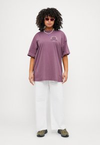 Purple oversized T-shirt with front graphic, paired with white wide-leg pants and olive green sneakers. Features curly hair and small sunglasses.
