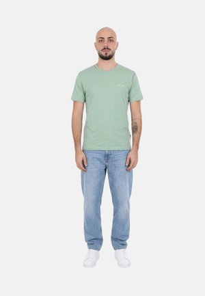 Uomo con avambraccio tatuato in piedi di fronte, indossa una t-shirt verde chiaro, jeans blu e sneakers bianche su uno sfondo uniforme.