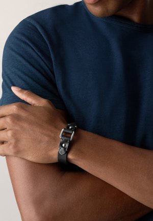 Bracelet en cuir noir avec quincaillerie en argent, présentant une boucle et des accents cloutés. Porté sur un bras bronzé, avec une chemise bleu marine.