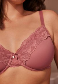 Mauve krajková podprsenka s hladkým materiálem a nastavovacími ramínky, s mašlí a malým ozdobným charmem ve středu.