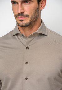 Camisa de un beige claro con una tela texturizada, que presenta un cuello abotonado y botones marrones. El diseño es elegante y ajustado, resaltando un aspecto moderno.