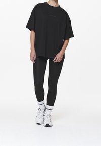 Černé oversize tričko z měkkého materiálu, nošené s černými legínami. Bílé ponožky s černým textem a bílé tenisky doplňují celkový outfit.