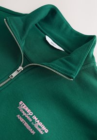 Grøn lynlåstrøje med ribstrikket krave. Har en sølvfarvet lynlås og pink broderet tekst, der lyder "STEREO IMAGING AMSTERDAM."