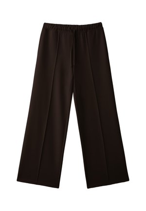 Femme debout les mains dans les poches, portant une chemise oversize à carreaux nouée à la taille, un pantalon ample foncé et une écharpe assortie à carreaux autour du cou.