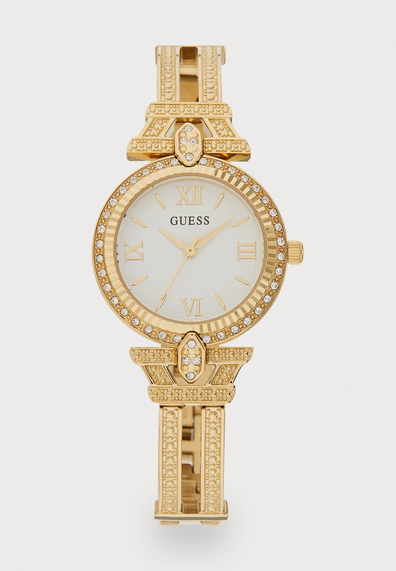 Montre dorée avec un cadran rond, un cadran blanc, des chiffres romains et des accents en cristal. Elle dispose d'un bracelet texturé avec un design orné.