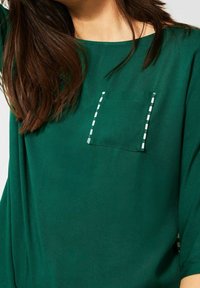 Zelená blůza s kulatým výstřihem, volným střihem a náprsní kapsou s bílými švy. Měkká tkanina s jemnou strukturou.