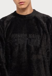 Μαύρη φλις μπλούζα από μαλακό ύφασμα, με ανάγλυφο λογότυπο "VERSACE JEANS COUTURE" σε σκούρο τόνο και στρογγυλή λαιμόκοψη.