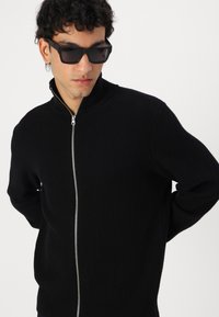 Čierny rebrovaný pletený sveter na zips s vysokým golierom a strieborným zipsom. Model nosí obrovské čierne slnečné okuliare, ruky má za chrbtom.