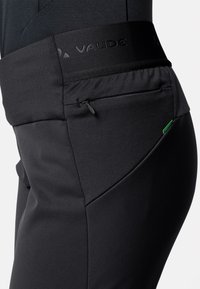 Pantalons de sport noirs avec une taille texturée, en matériau élastique, et une poche zippée sur le côté. Présente une étiquette d'accent vert.