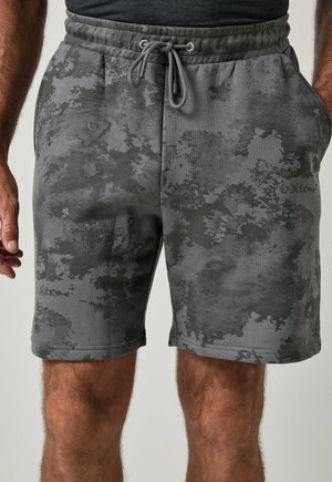 Uomo che indossa pantaloncini grigi con motivo mimetico e coulisse, mano sinistra in tasca e mano destra rilassata di fianco.