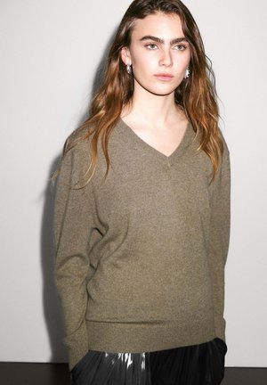 Maglione verde oliva a scollo a V in morbido tessuto lavorato a maglia, con polsini e orlo a costine. Abbinato a una gonna plissettata nera lucida.