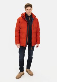 Rød isoleret pufferjakke med hætte, lynlåslommer og en frontlynlås. Båret over en sort top, parret med mørke jeans og beige støvler.
