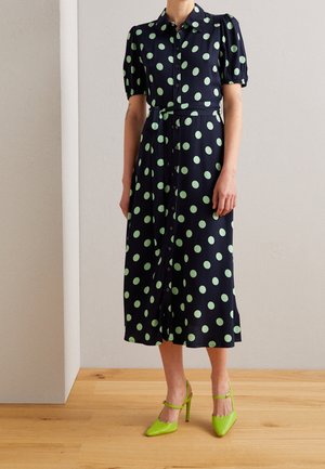 Vrouw draagt een marineblauwe midi-jurk met grote groene polkadots, pofmouwen, knopen vooraan, tailleceintuur en limoengroene hoge hakken.