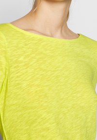 Haut en tricot léger de couleur jaune vif avec un décolleté large et des manches courtes. Le tissu a une apparence légèrement texturée et transparente.
