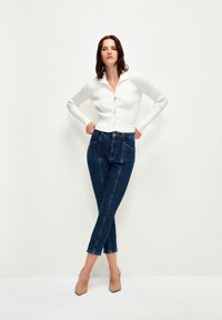 Bílý žebrovaný cardigan s knoflíky, který je spojen s tmavými džíny s vysokým pasem a švy. Oblečené s nahými podpatky.