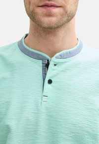 Světle zelená texturovaná košile s kulatým límcem, s šedým vnitřním límcem, dvěma černými knoflíky a minimalistickými švy.