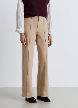 Donna che indossa pantaloni beige a gamba larga, camicia bianca a maniche lunghe con polsini arrotolati, maglione bordeaux senza maniche e mocassini neri.