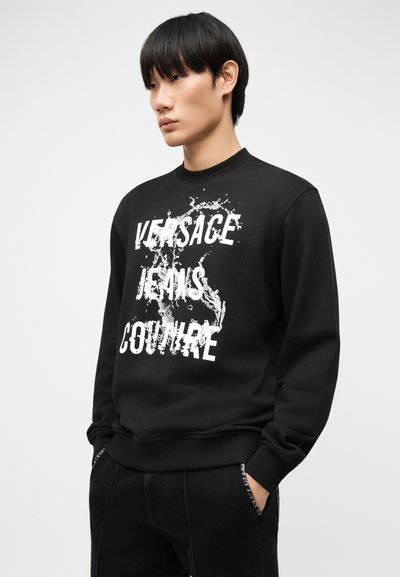 Jeune homme portant un sweat-shirt noir avec le texte blanc "Versace Jeans Couture" et un motif d'éclaboussures de peinture, mains dans les poches, fond neutre.