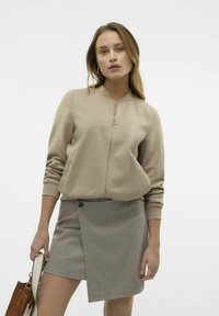 Veste bomber beige zippée avec poches, associée à une jupe enveloppante à motif pied-de-coq avec un détail de bouton.