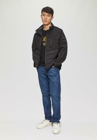 Zwarte gewatteerde jas met ritszak, grijze accenten, gelaagd ontwerp; gecombineerd met een zwart shirt, blauwe jeans en witte sneakers.