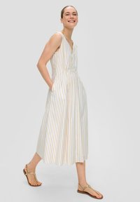 Robe midi sans manches rayée en blanc avec des rayures dorées et bleues. Dotée d'un décolleté en V, d'une taille cintrée et de poches latérales. Portée avec des sandales beiges.