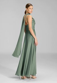 Abito lungo in raso verde fluente con schiena drappeggiata, spalline sottili e trama morbida, abbinato a scarpe con tacco alto argento.