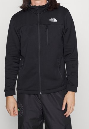 Osoba ubrana w czarną kurtkę zapinaną na zamek marki The North Face z kieszeniami na zamek oraz czarne spodnie sportowe z zielonym logo.